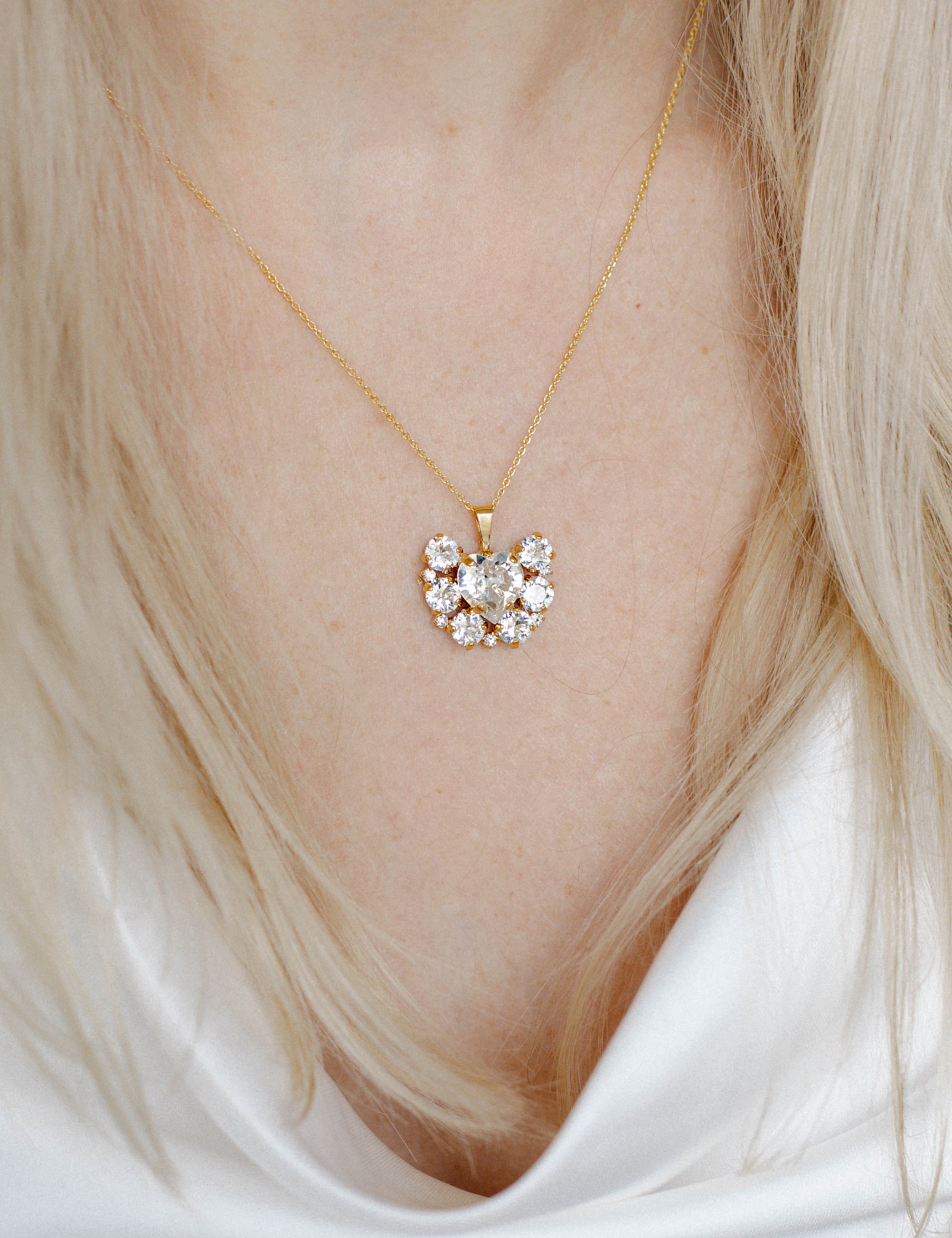 A person wearing a white v-neck top showcasing a gold-plated necklace with a floral pattern and a central heart-shaped crystal pendant.