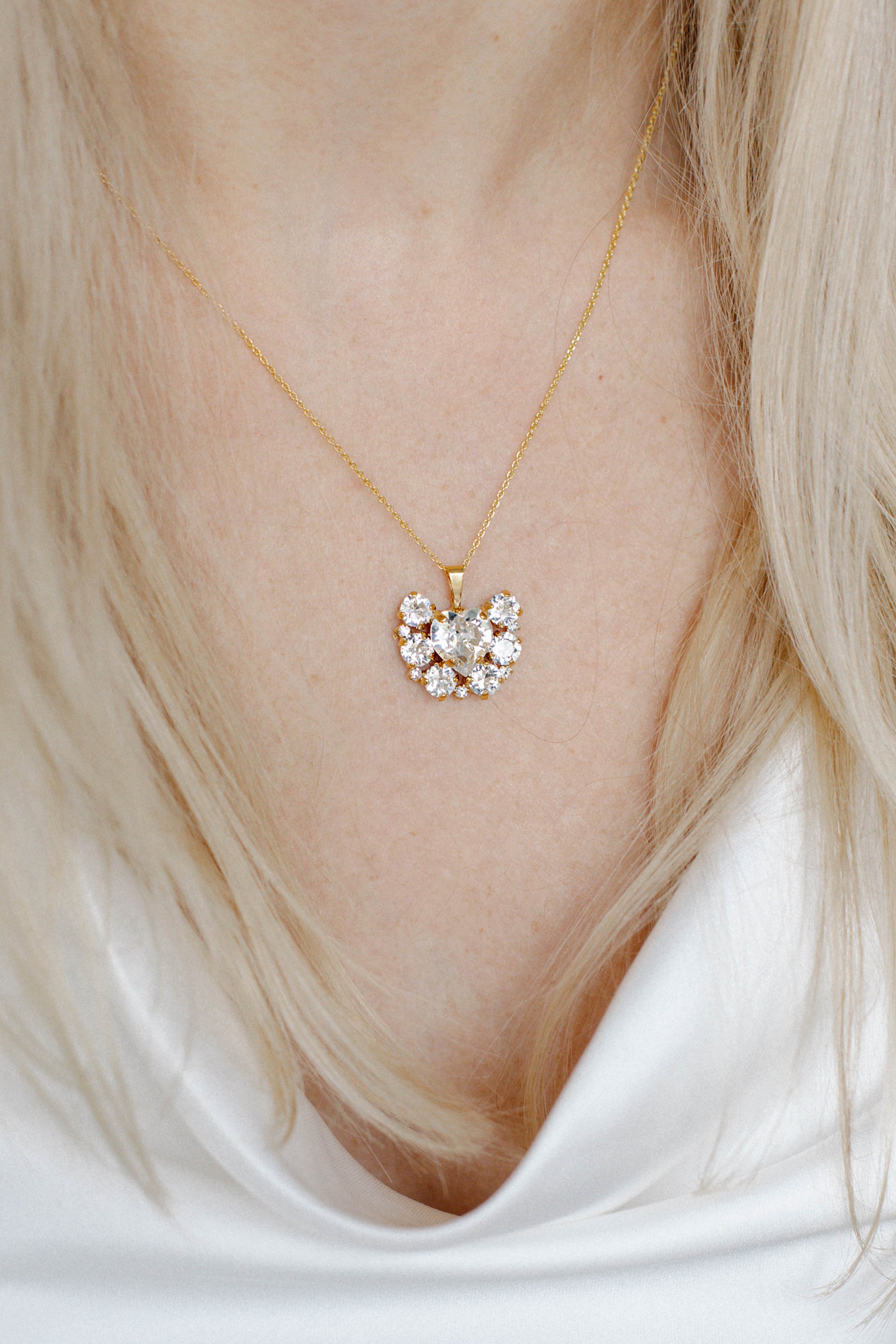 A person wearing a white v-neck top showcasing a gold-plated necklace with a floral pattern and a central heart-shaped crystal pendant.