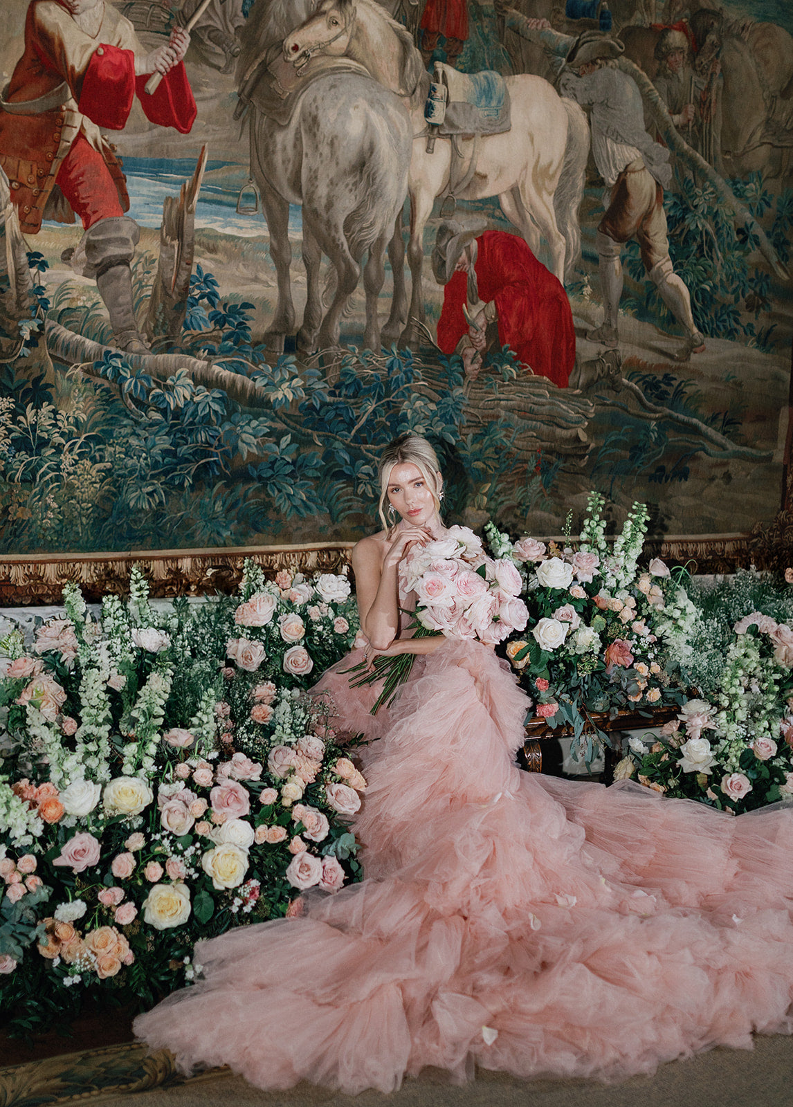 Woman in a pink dress sitting among flowers with a tapestry in the background
