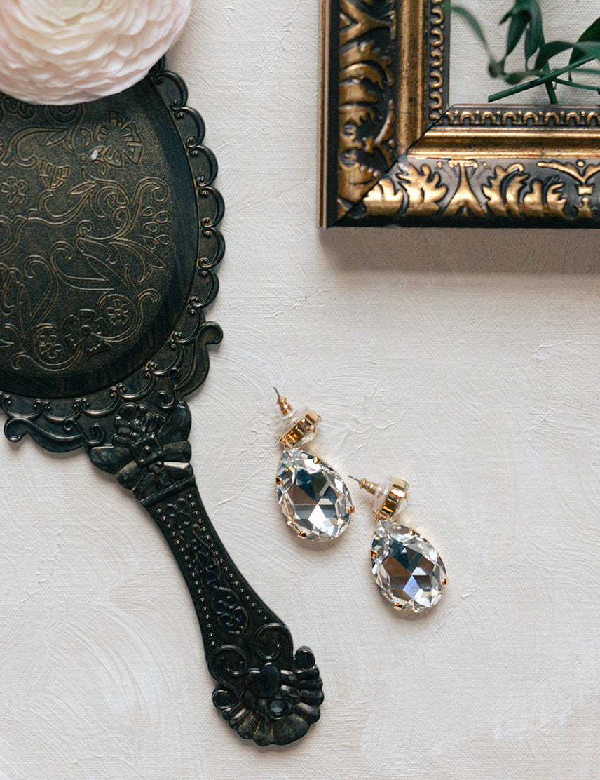 Vintage-style hand mirror, pink flower, and crystal earrings on a light surface.