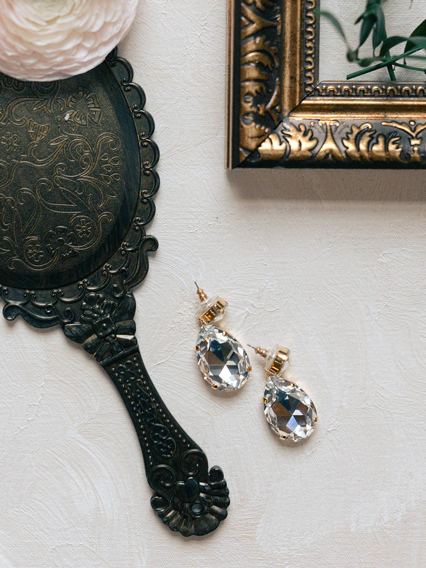 Vintage-style hand mirror, pink flower, and crystal earrings on a light surface.