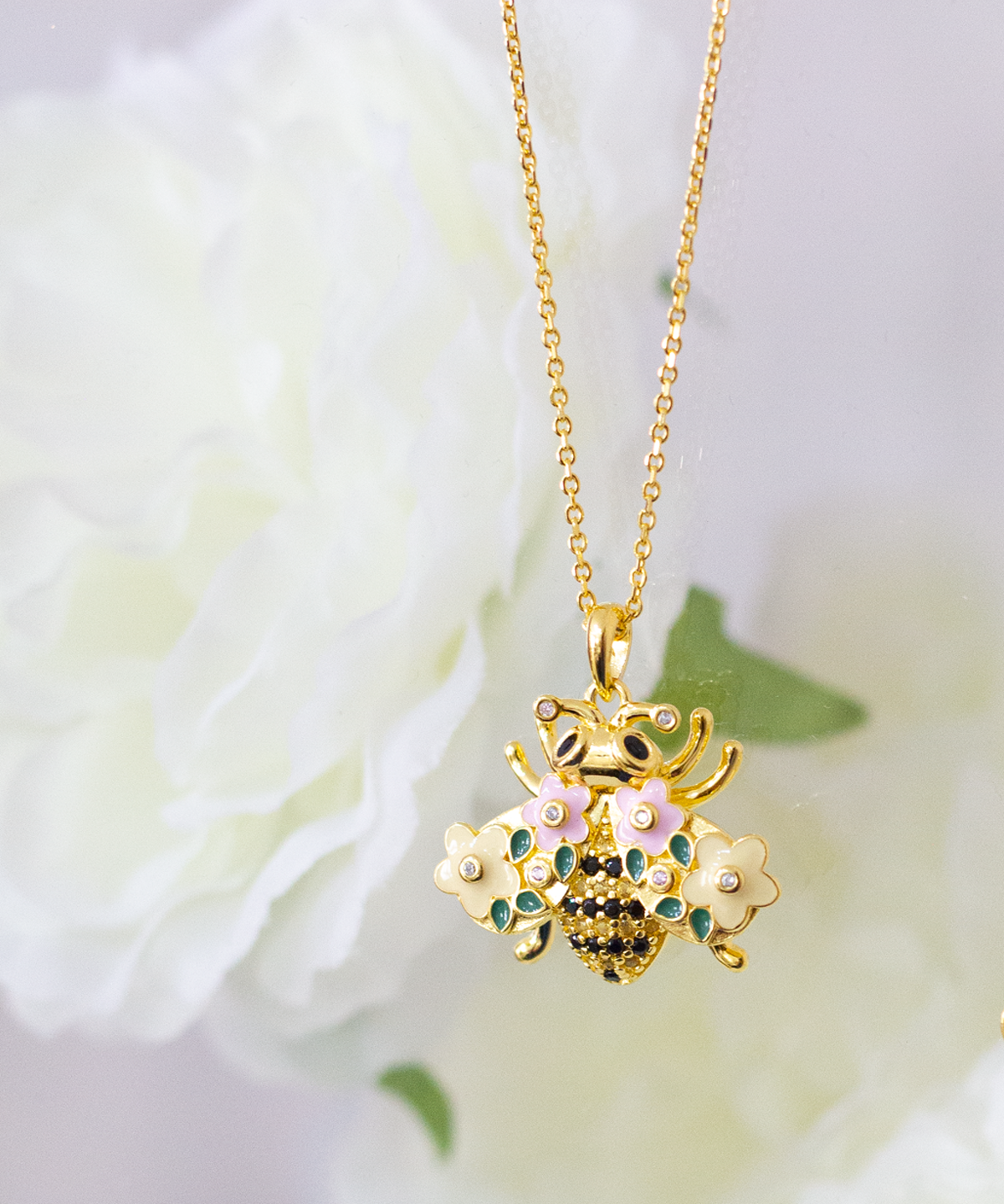 A gold-plated bumble bee necklace with a chain and a floral pattern, displayed against a soft-focus background with flowers.