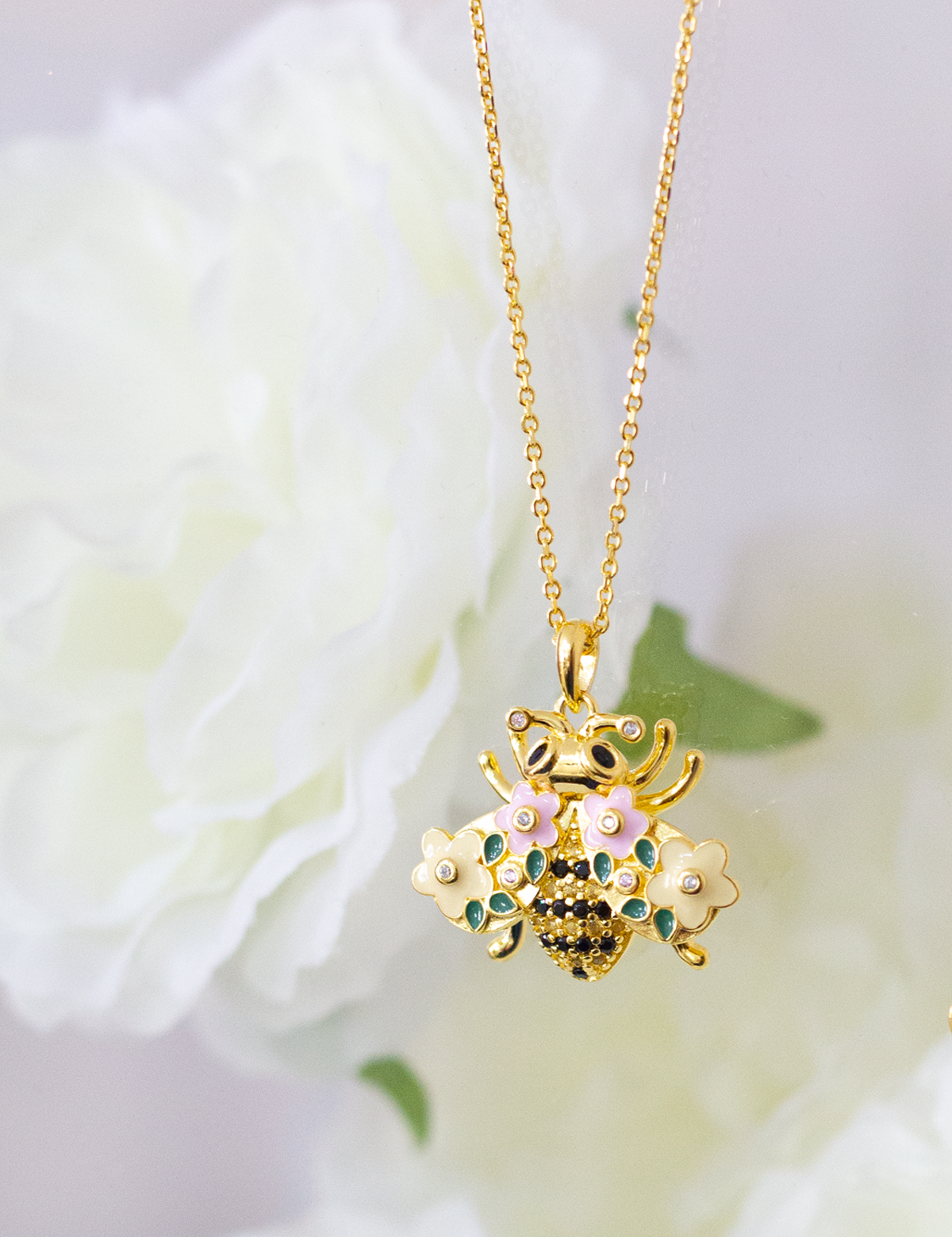 A gold-plated bumble bee necklace with a chain and a floral pattern, displayed against a soft-focus background with flowers.