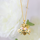 A gold-plated bumble bee necklace with a chain and a floral pattern, displayed against a soft-focus background with flowers.