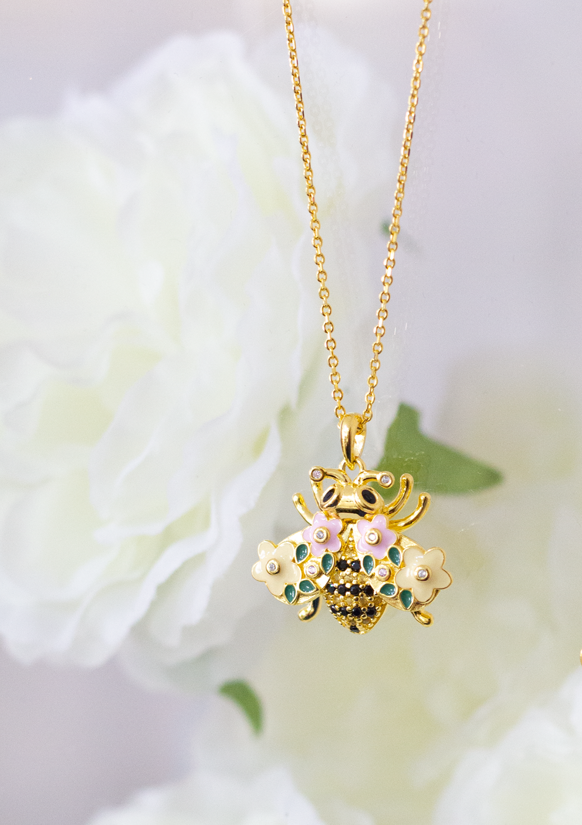 A gold-plated bumble bee necklace with a chain and a floral pattern, displayed against a soft-focus background with flowers.