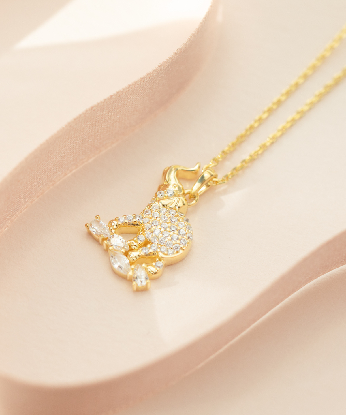 A gold-plated sterling silver elephant pendant necklace with a cubic zirconia crystal on top, accompanied by a matching chain and extender, displayed on a pink surface.