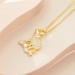 A gold-plated sterling silver elephant pendant necklace with a cubic zirconia crystal on top, accompanied by a matching chain and extender, displayed on a pink surface.