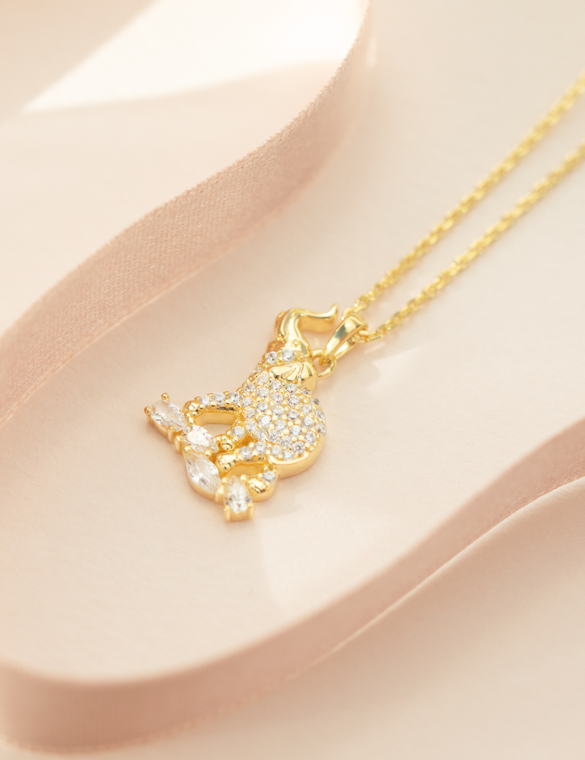 A gold-plated sterling silver elephant pendant necklace with a cubic zirconia crystal on top, accompanied by a matching chain and extender, displayed on a pink surface.