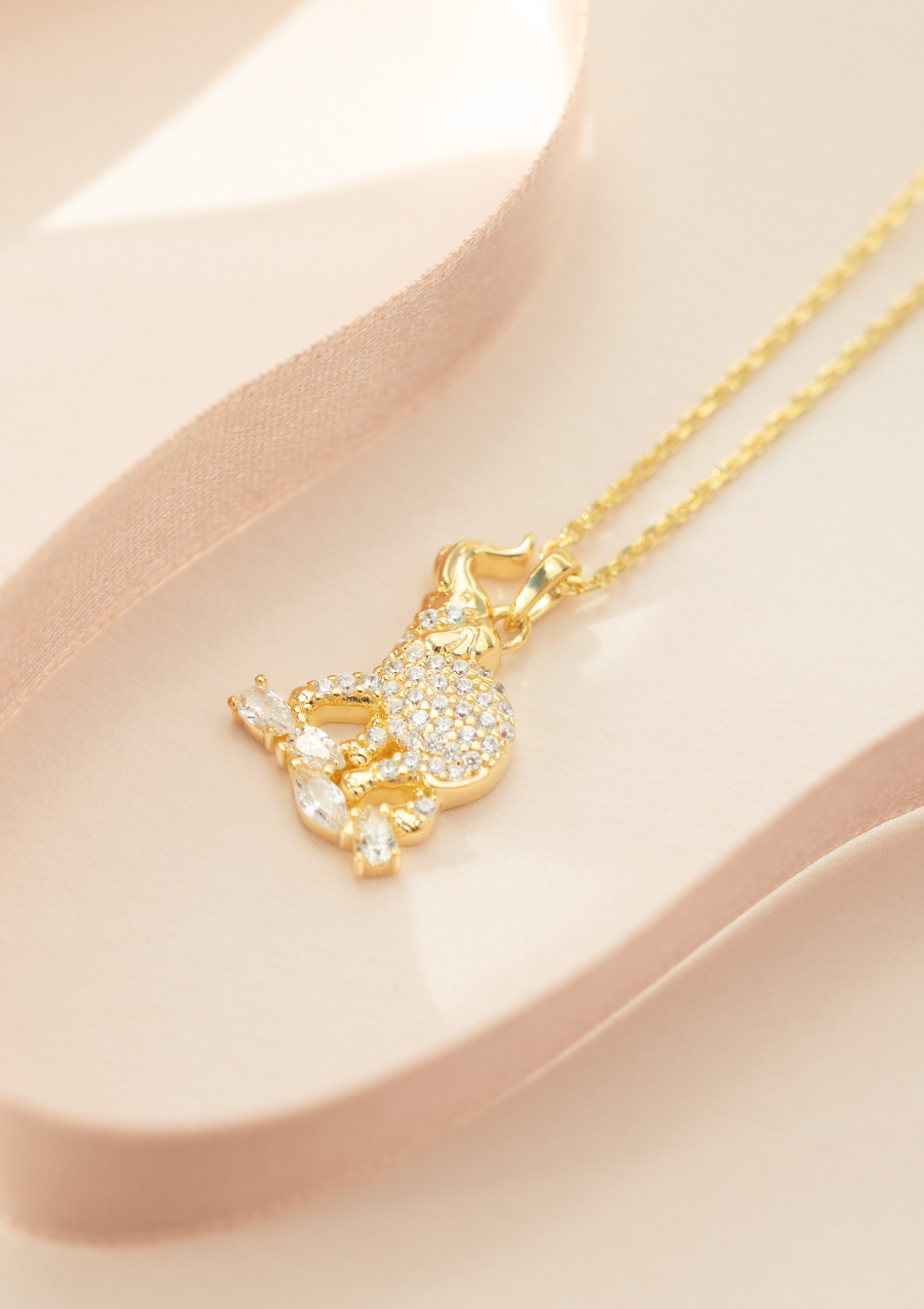 A gold-plated sterling silver elephant pendant necklace with a cubic zirconia crystal on top, accompanied by a matching chain and extender, displayed on a pink surface.