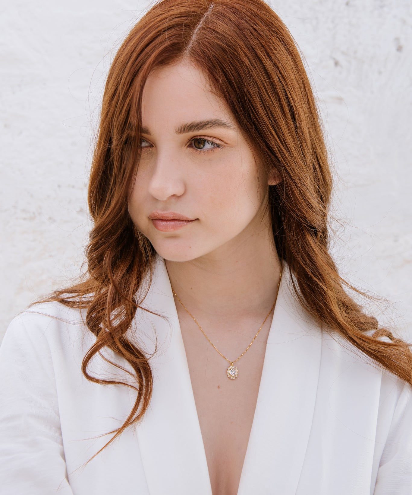 A woman with red hair wearing a white blazer, featuring a duchess necklace with a central oval crystal surrounded by smaller crystals.