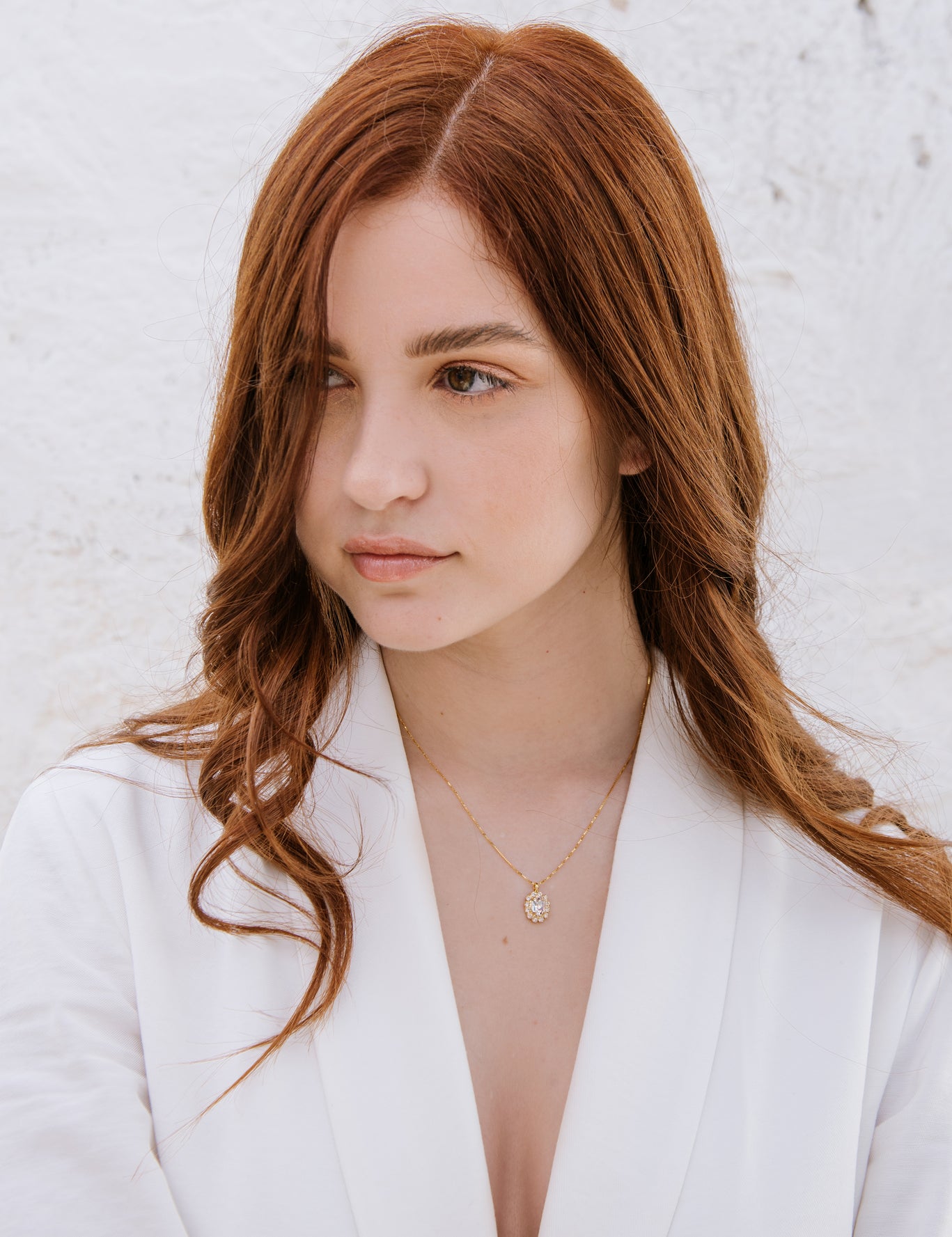 A woman with red hair wearing a white blazer, featuring a duchess necklace with a central oval crystal surrounded by smaller crystals.