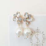A pair of gold-colored earrings featuring baroque pearls and sparkling crystals, placed on a white card with the logo 'SARAH GAUCI'.