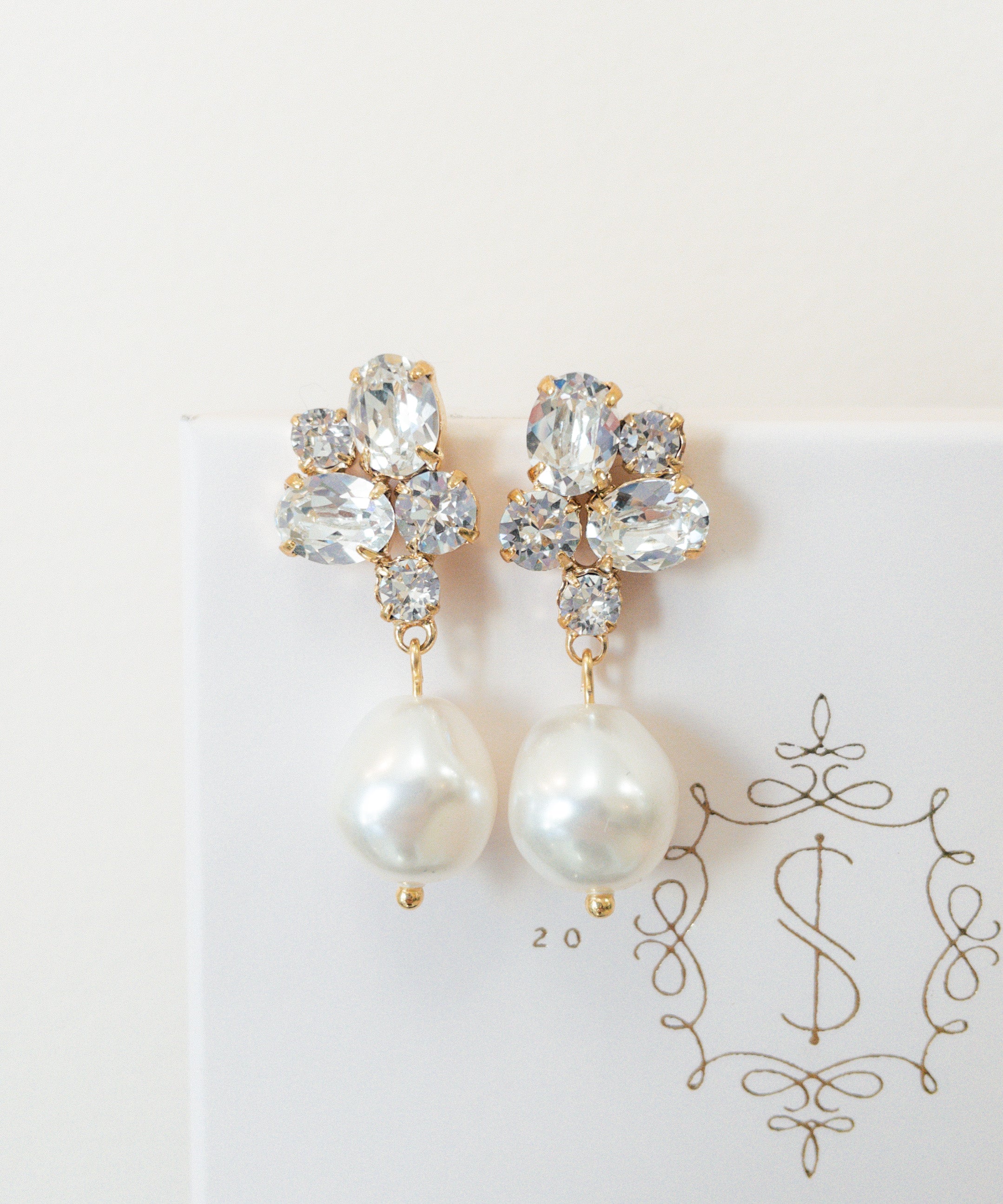 A pair of gold-colored earrings featuring baroque pearls and sparkling crystals, placed on a white card with the logo 'SARAH GAUCI'.