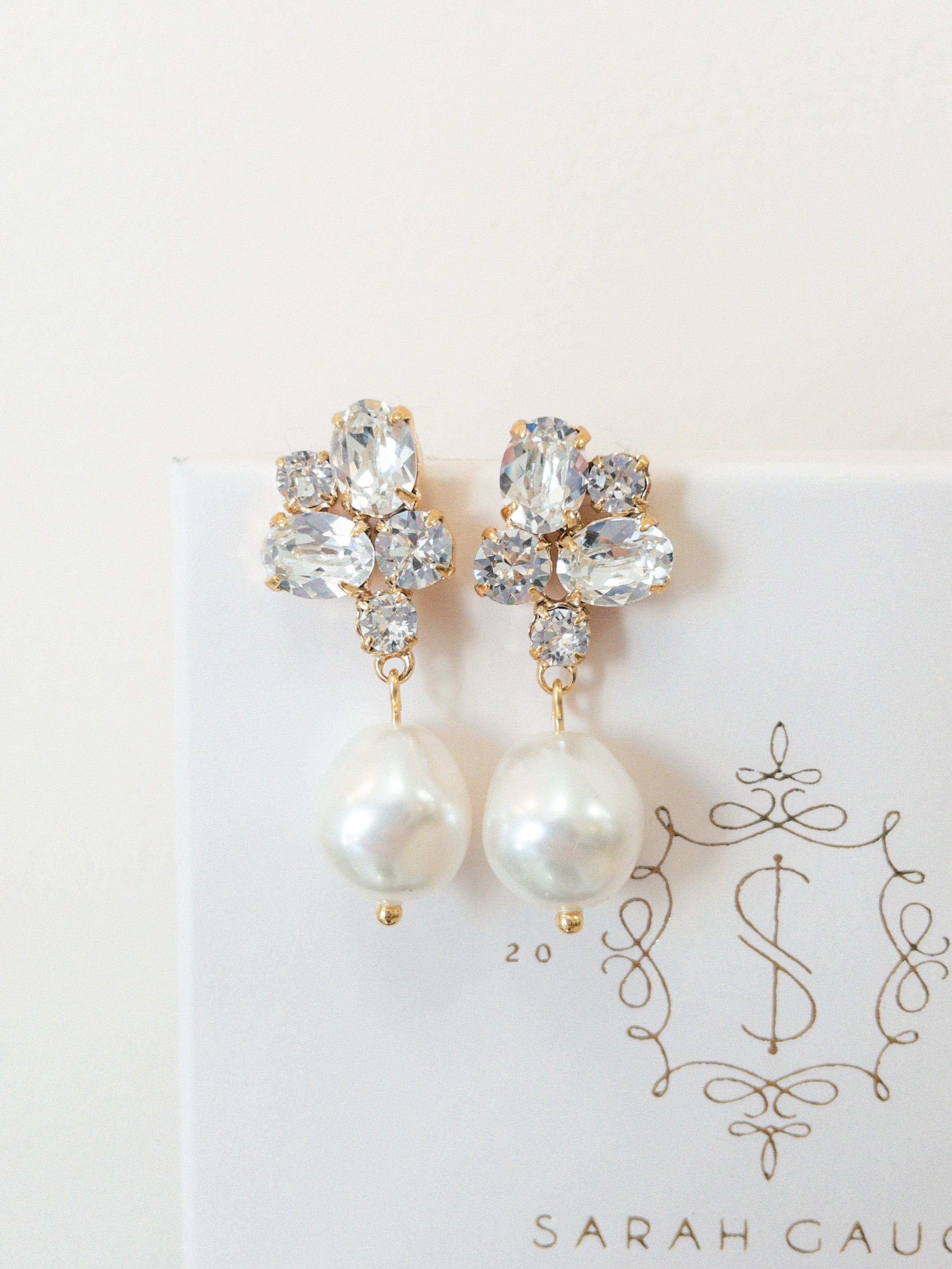 A pair of gold-colored earrings featuring baroque pearls and sparkling crystals, placed on a white card with the logo 'SARAH GAUCI'.