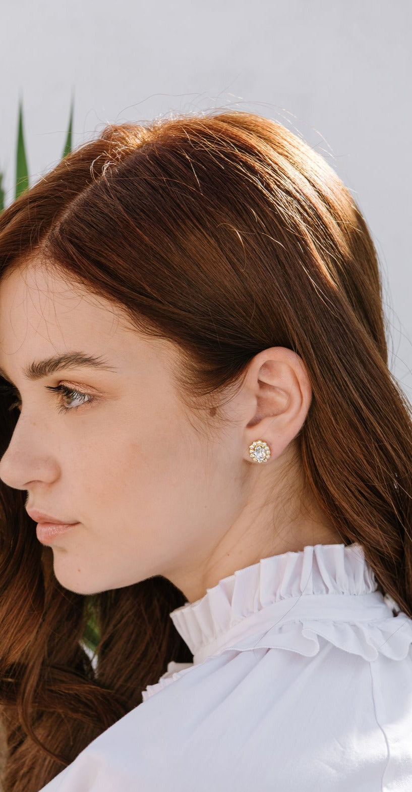 A woman wearing a white top with a crew neckline, showcasing emerald green earrings surrounded by clear crystals.