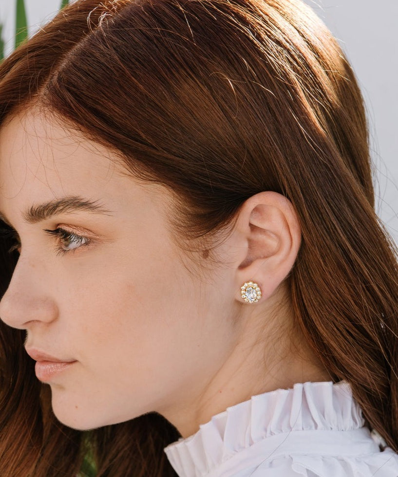 A woman wearing a white top with a crew neckline, showcasing emerald green earrings surrounded by clear crystals.