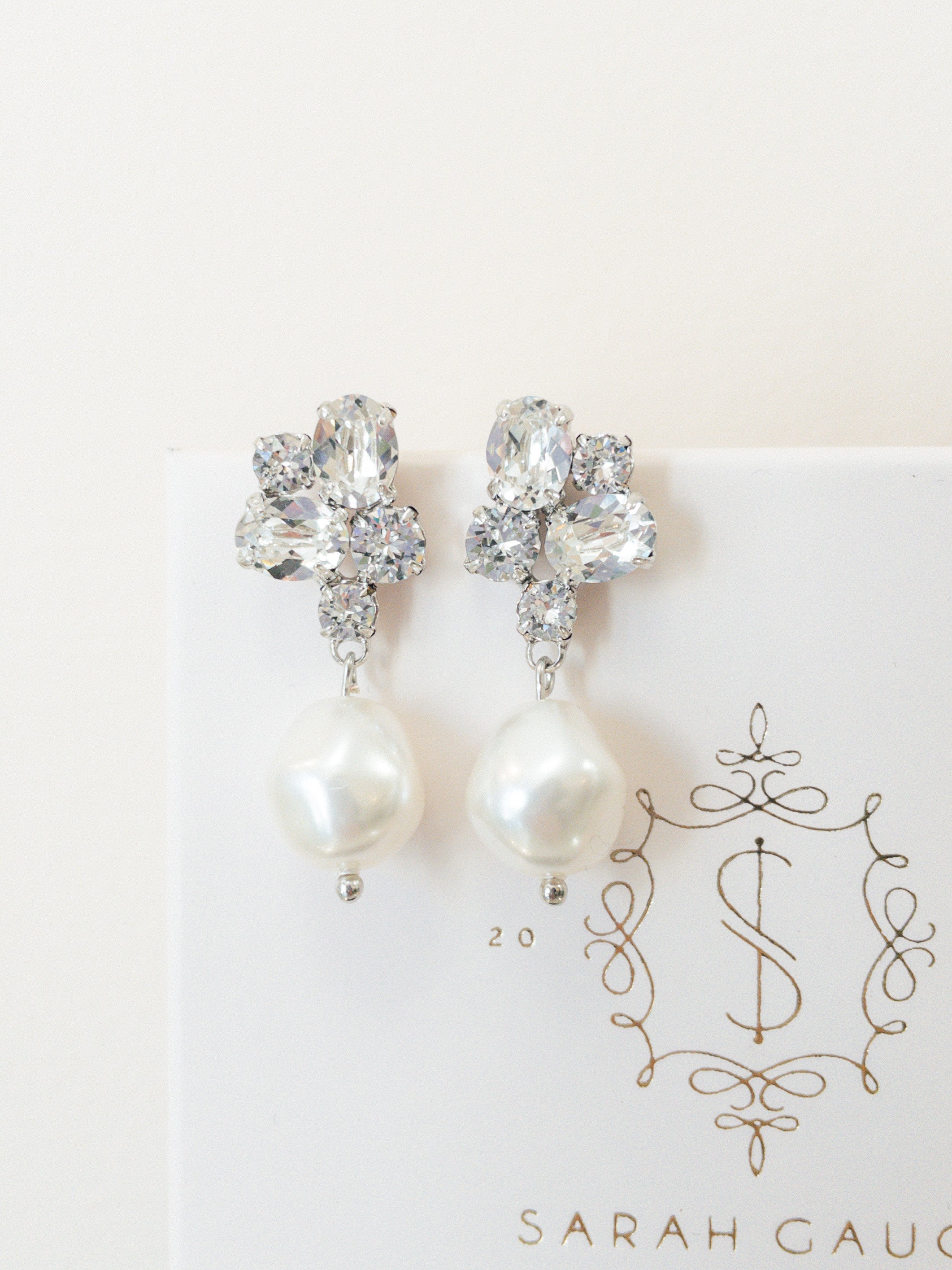 A pair of silver earrings featuring baroque pearls and sparkling crystals, placed on a white card with the logo 'SARAH GAUCI'.