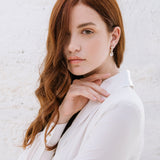 A woman with red hair wearing a white shirt, showcasing the Brianna earrings which are hanging from her ear. The earrings are shiny with a rhodium or gold plating and adorned with high-quality crystals.