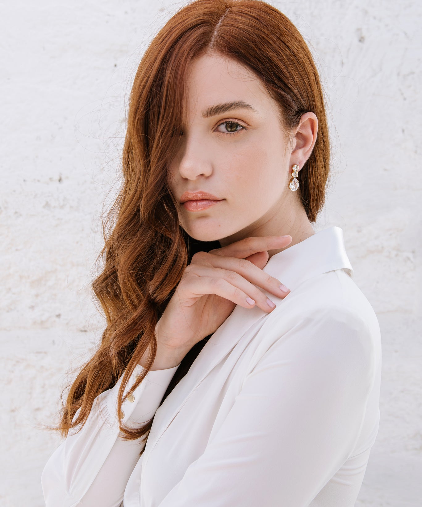 A woman with red hair wearing a white shirt, showcasing the Brianna earrings which are hanging from her ear. The earrings are shiny with a rhodium or gold plating and adorned with high-quality crystals.