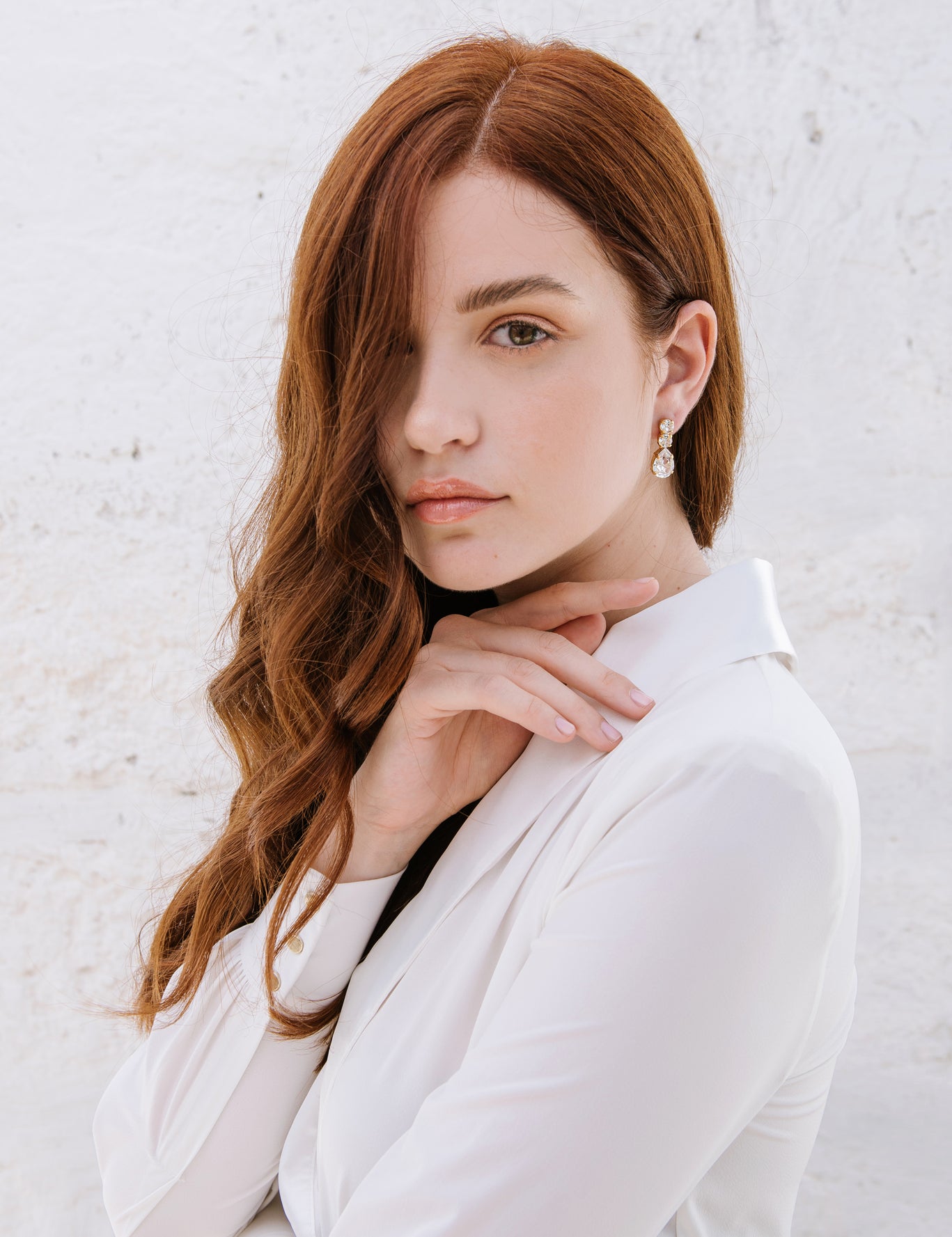 A woman with red hair wearing a white shirt, showcasing the Brianna earrings which are hanging from her ear. The earrings are shiny with a rhodium or gold plating and adorned with high-quality crystals.