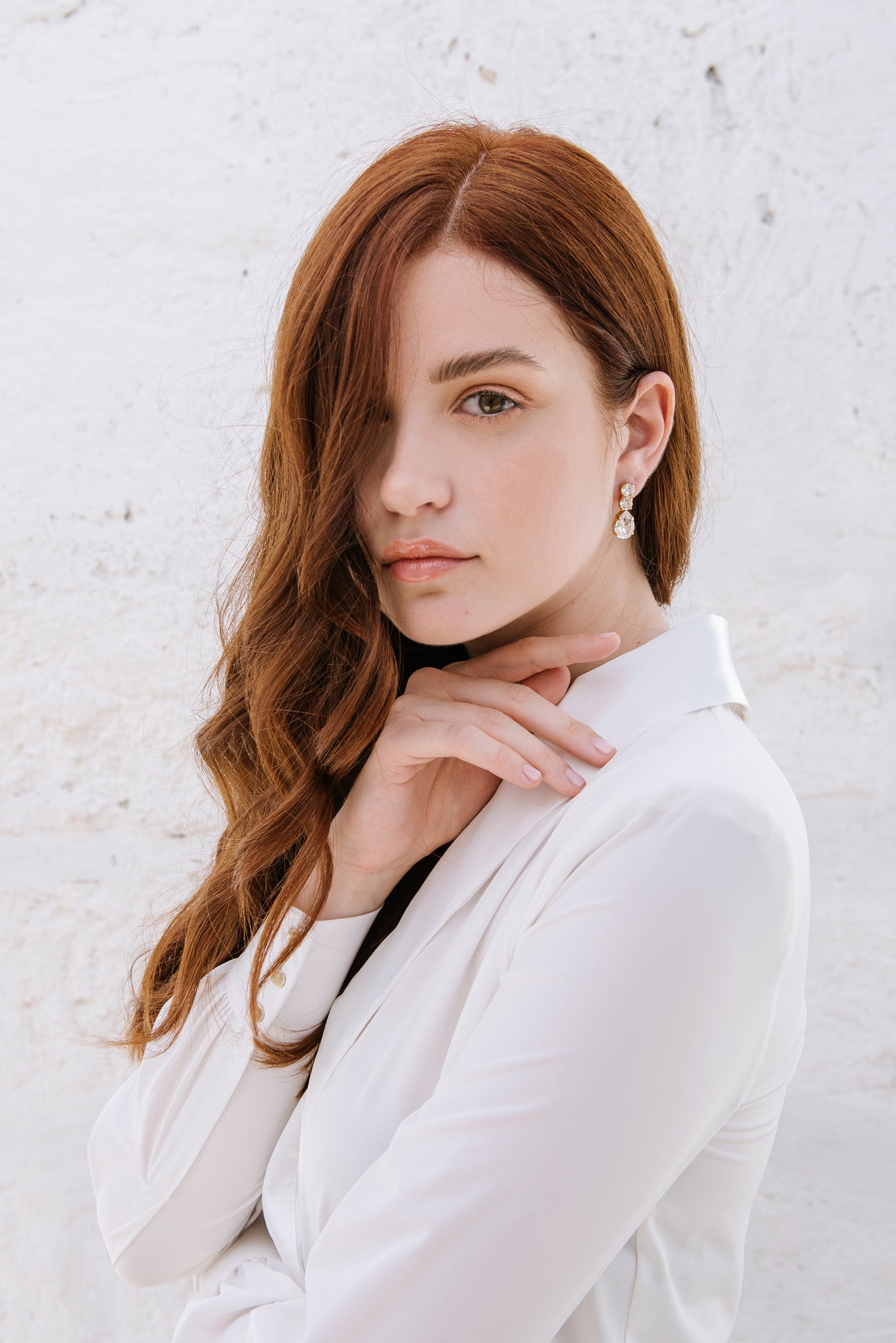 A woman with red hair wearing a white shirt, showcasing the Brianna earrings which are hanging from her ear. The earrings are shiny with a rhodium or gold plating and adorned with high-quality crystals.